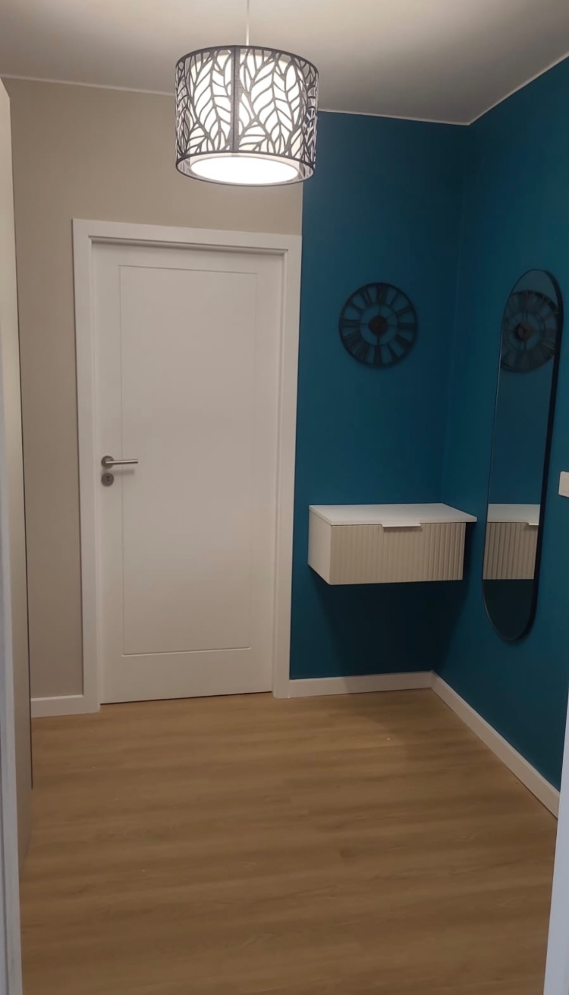 Nowoczesny przedpokój: białe drzwi, szafka wisząca, lustro w czarnej ramie na tle turkusowej ściany. Lampa sufitowa z motywem liści. Jasna podłoga.