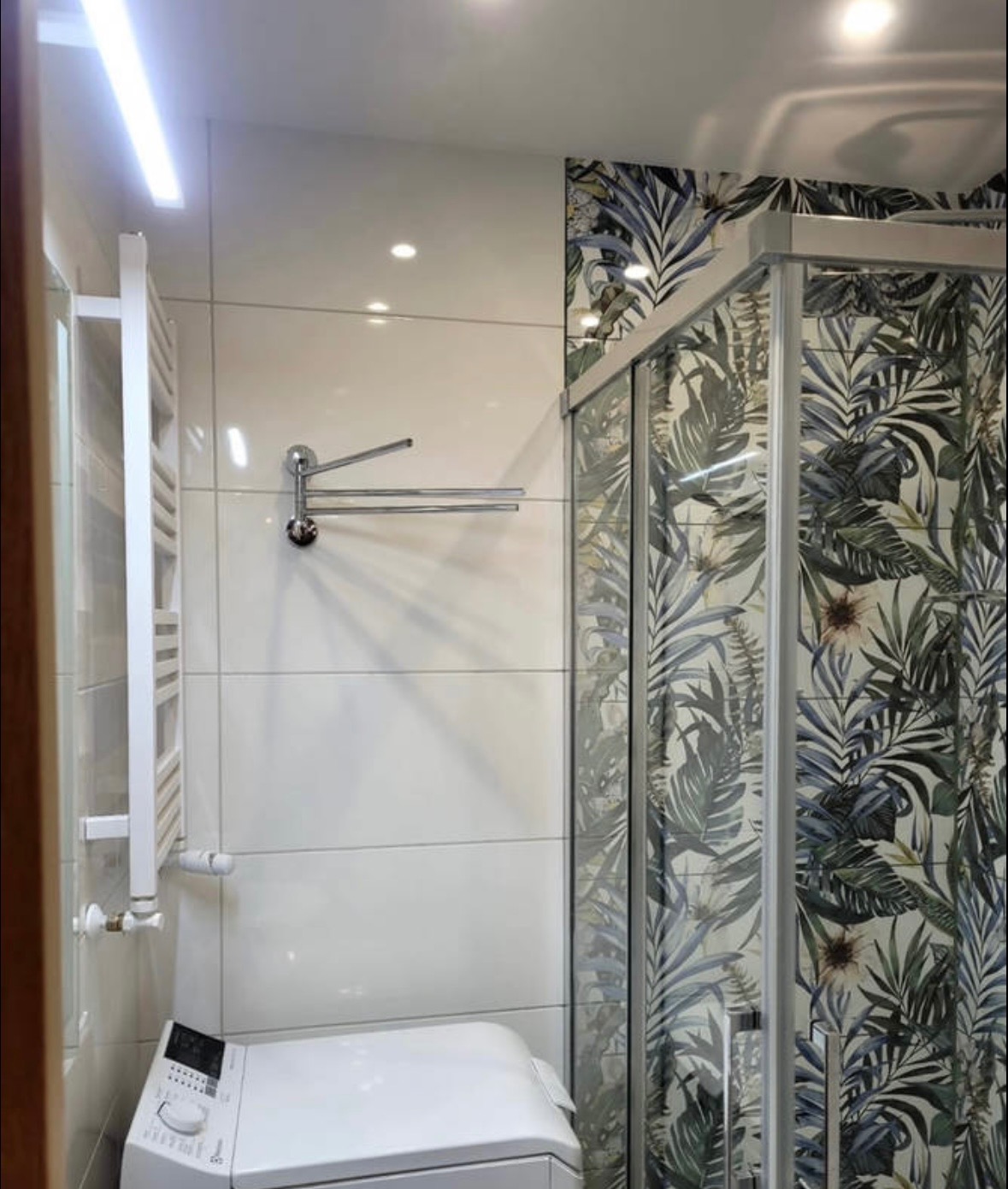 Wykończona łazienka z kabiną prysznicową w tropikalne wzory, białą pralką i grzejnikiem drabinkowym. Oświetlenie LED i chromowane akcesoria.