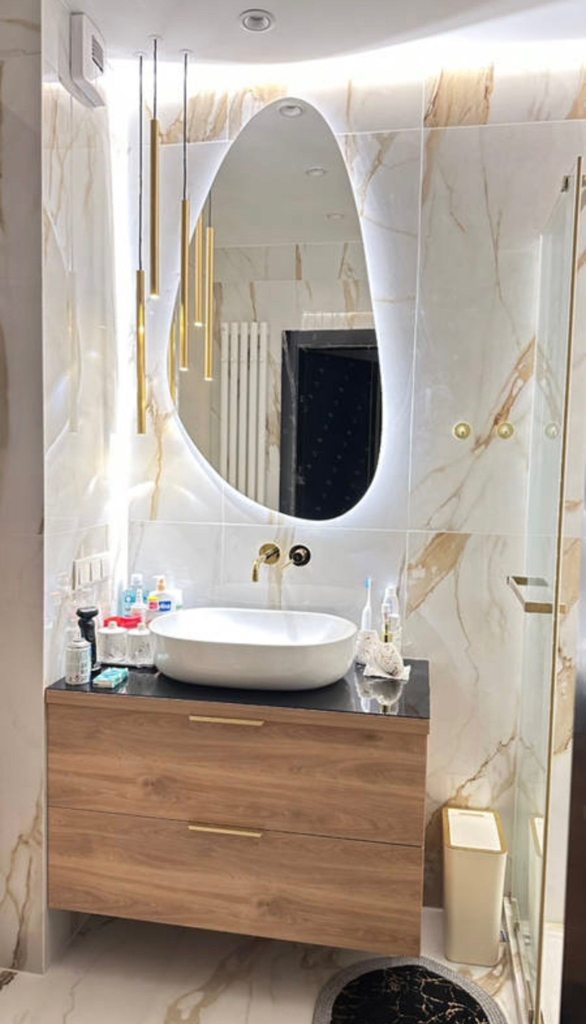 Elegancka łazienka z owalnym lustrem podświetlanym LED, wiszącą szafką z umywalką i złotymi lampami. Marmurowe płytki na ścianach i podłodze.