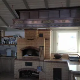 Wnętrze kuchni w domu z bali z ceglanym piecem chlebowym, drewnianymi szafkami i dekoracyjnym okapem wentylacyjnym.