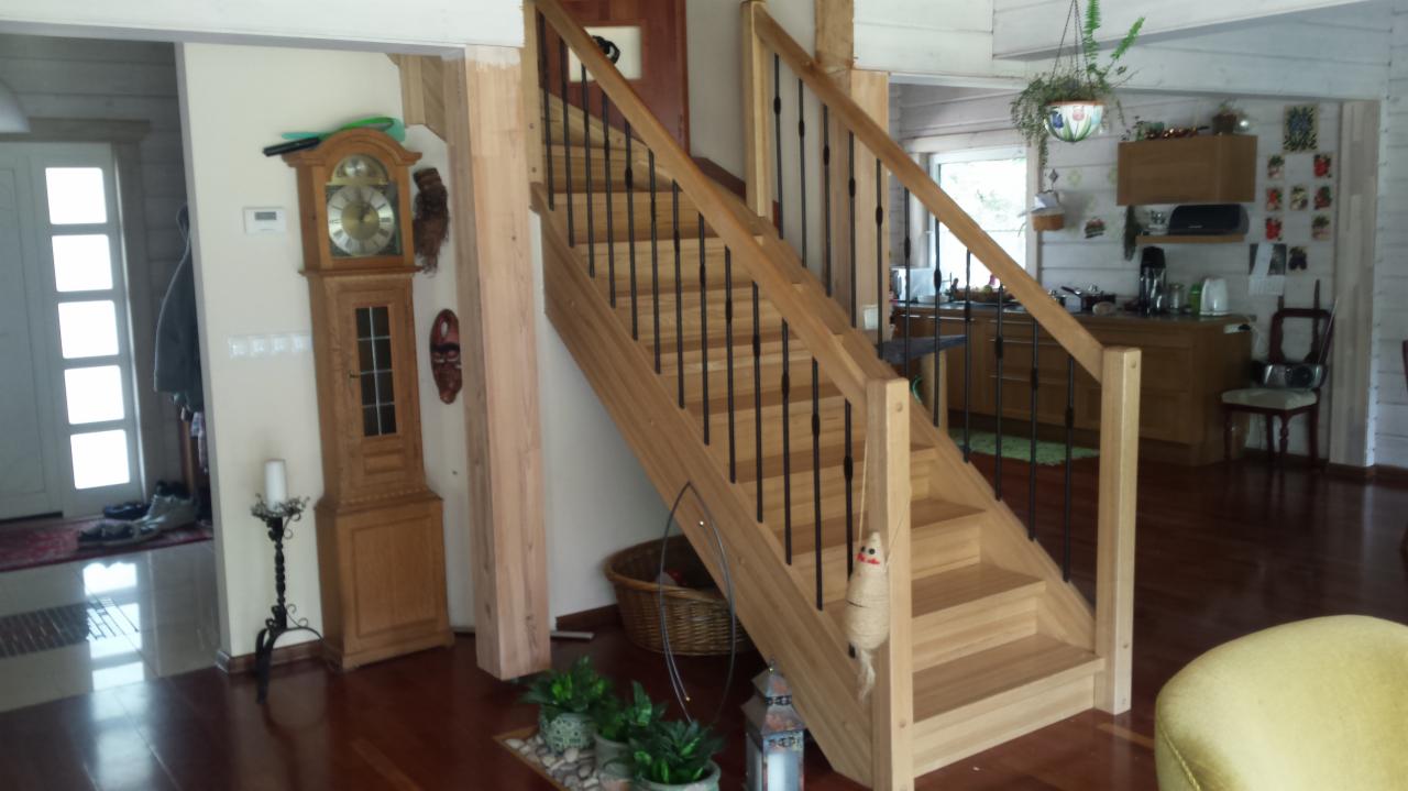 Wnętrze domu z drewnianymi schodami z czarnymi balustradami, widoczny zegar stojący, dekoracje roślinne i fragment kuchni w tle.