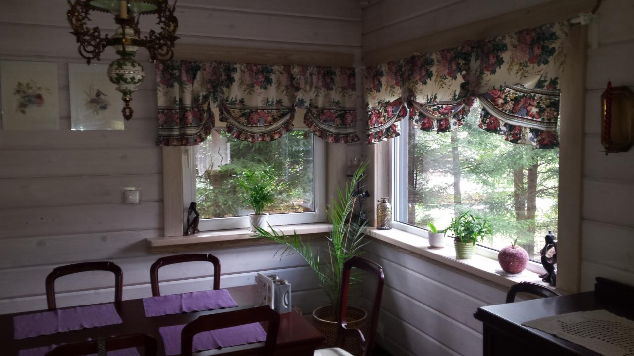 Wnętrze pokoju w domu z drewnianych bali, z widokiem na las przez okna ozdobione kwiatowymi lambrekinami, drewniany stół z fioletowymi podkładkami i krzesłami, rośliny doniczkowe na parapecie.