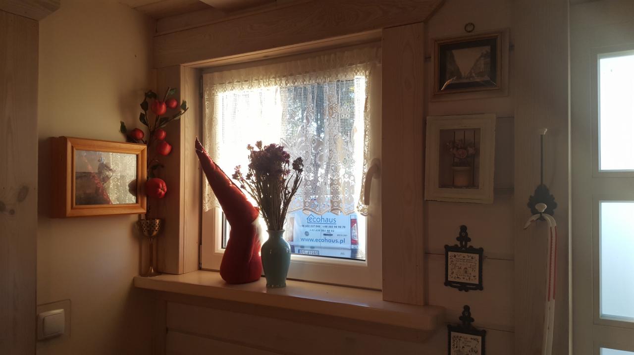 Jasne wnętrze domu z drewnianym oknem, parapetem z dekoracjami: wazon z kwiatami, czerwona czapka elfa, jabłka dekoracyjne, na ścianie wiszą obrazki w drewnianych ramkach.