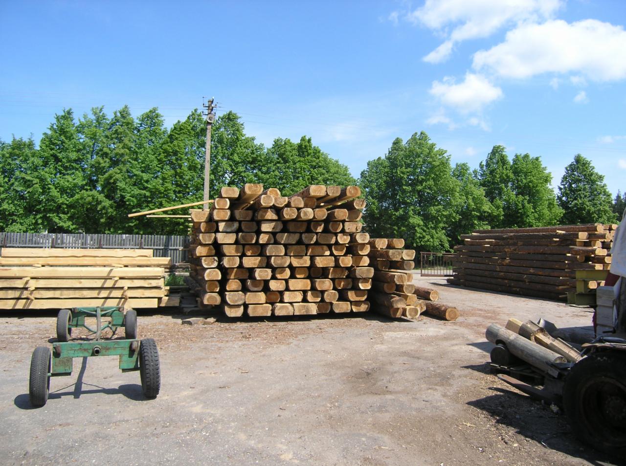 Stosy obrobionych bali drewnianych na placu składowym, z wózkiem transportowym i fragmentem ogrodzenia w tle.