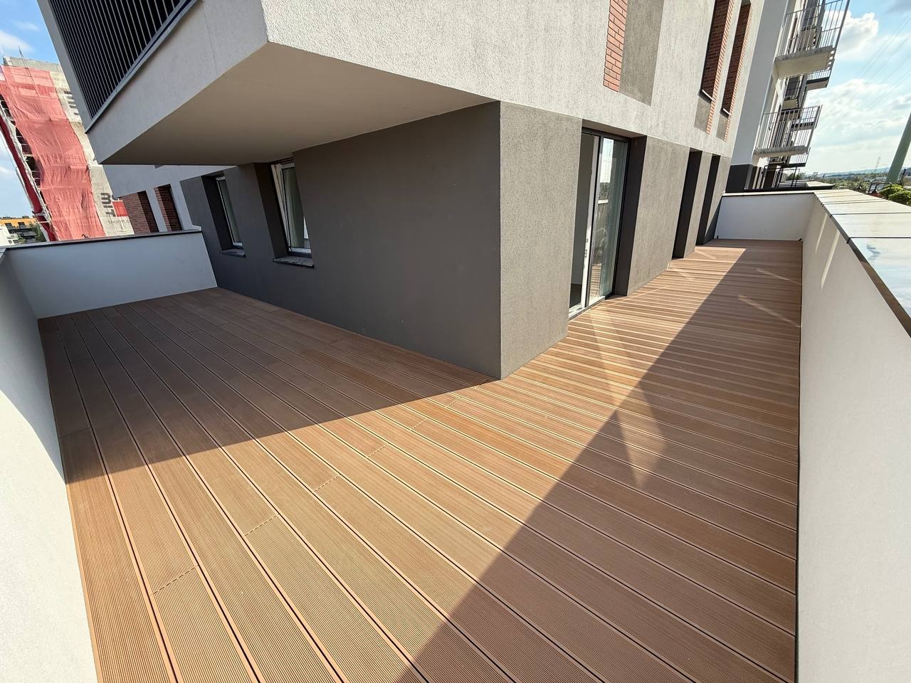 Nowoczesny balkon z deską kompozytową w odcieniach brązu, z widokiem na miasto w tle. Minimalistyczna balustrada i gra cieni podkreślają estetykę.