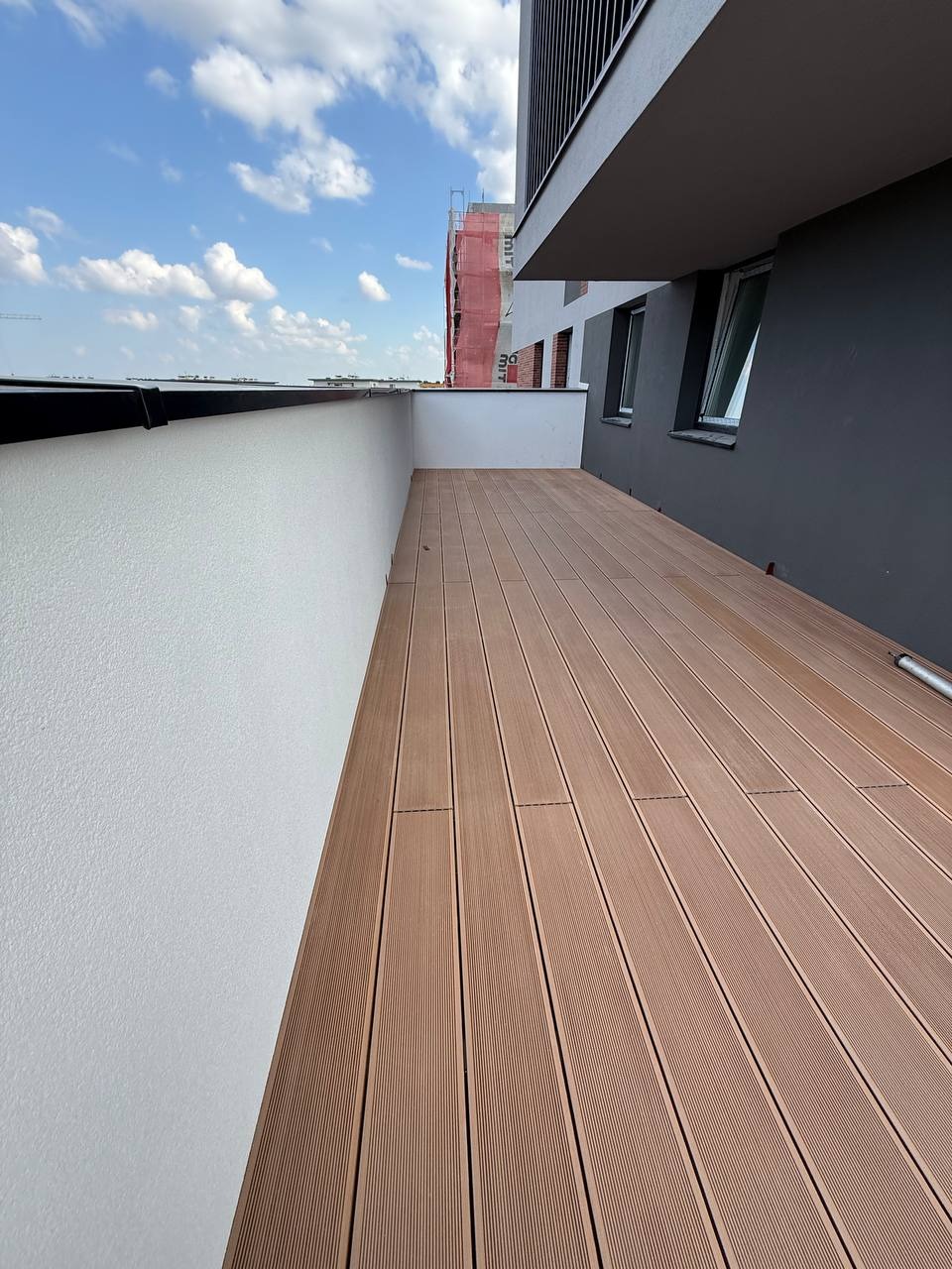 Wykończony balkon z brązową deską kompozytową, białą ścianą i widokiem na budynek w tle. Nowoczesny design i estetyczne wykończenie.