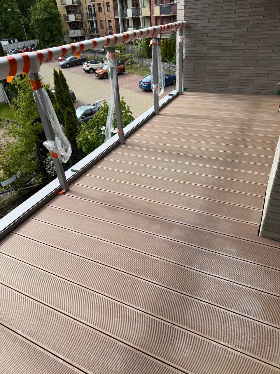 Balkon z deskami kompozytowymi, szara balustrada z folią ochronną, widok na parking i budynek. Deski ułożone równolegle. Nowoczesny wygląd.