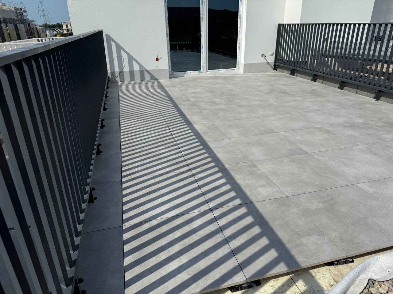 Wykończony balkon z szarymi płytkami, widoczne czarne barierki i cień. Nowoczesne wykończenie z dbałością o detale.