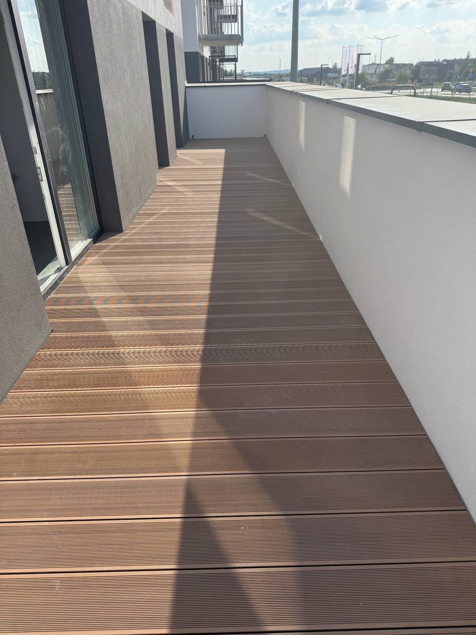 Wykończony balkon z brązową deską kompozytową ułożoną wzdłuż, jasne ściany i szara elewacja budynku w tle, widok na miasto w oddali.