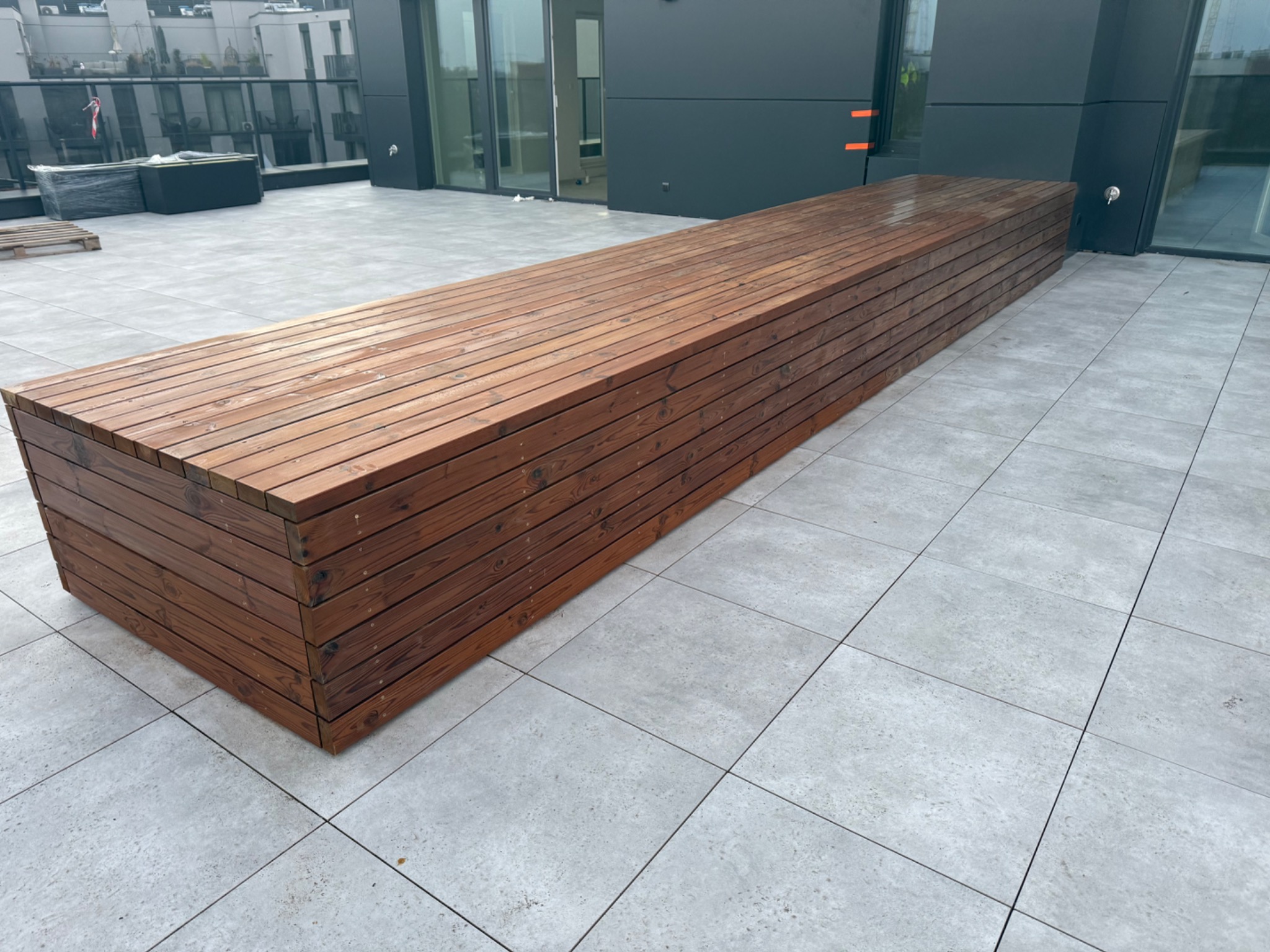 Długi, drewniany siedzisko/ławka na szarym tarasie z płytek. Nowoczesny design, minimalistyczne otoczenie, widok na budynek z przeszklonymi balkonami.