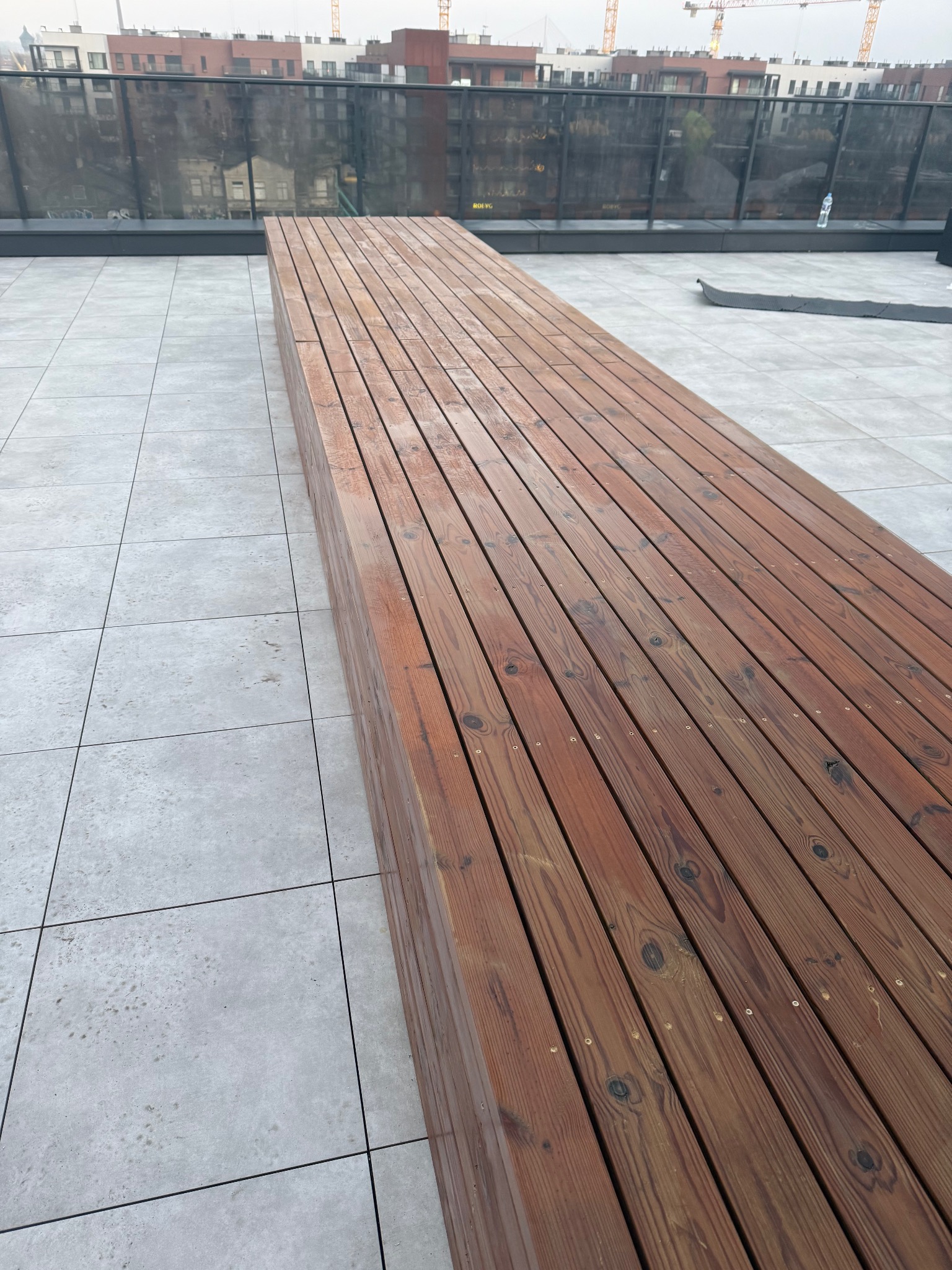 Długi, drewniany podest z desek na szarych płytkach tarasu. W tle budynki i żurawie budowlane. Widok z góry, lekki kąt.