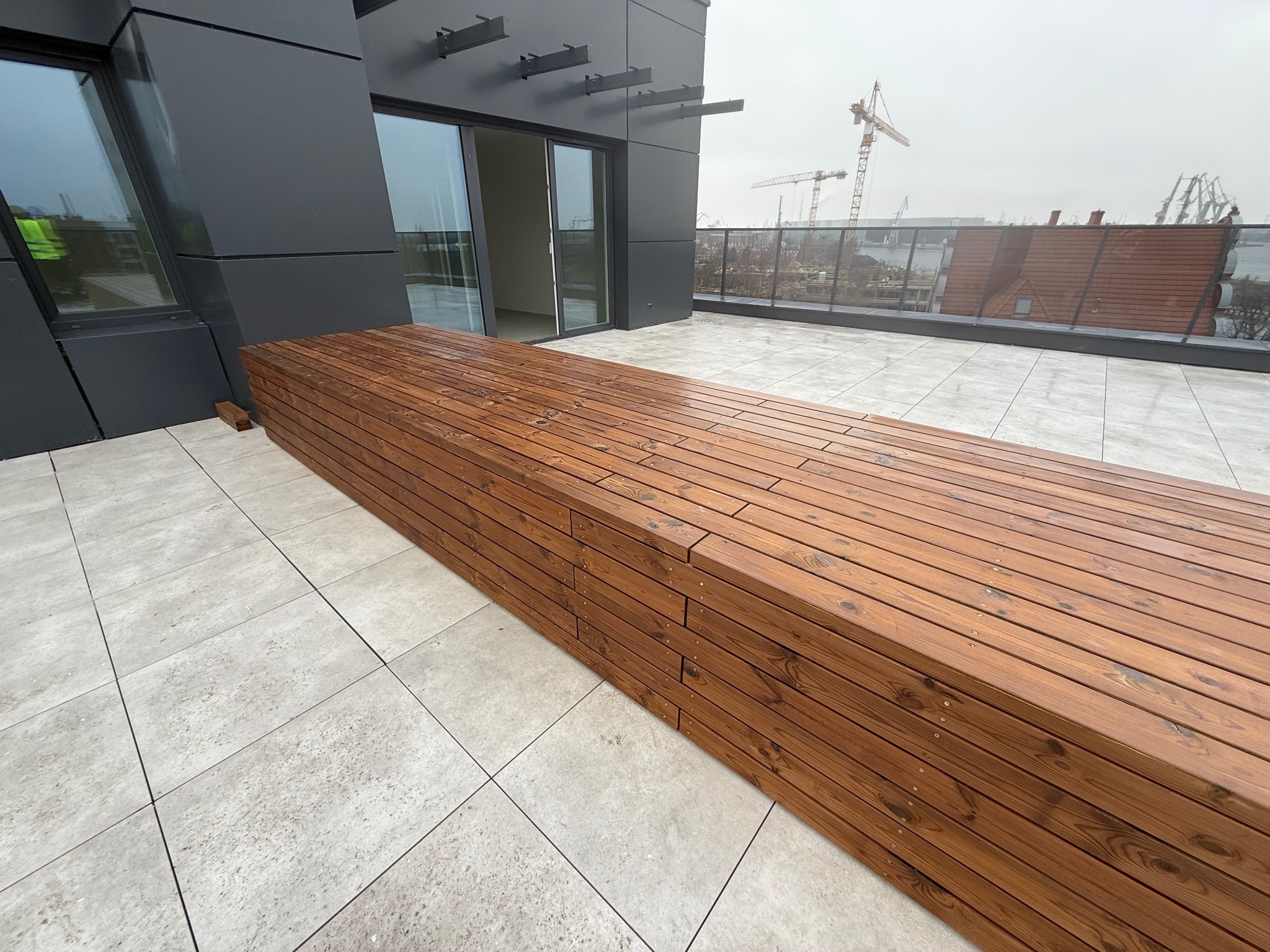 Nowoczesny taras w Gdańsku z drewnianą ławką i szarą płytką. Widok na portowe dźwigi w tle. Minimalistyczny design, deski ułożone poziomo.