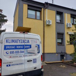 OPTYMAL SYSTEM RAFAŁ KONDEJ - Żółto-szary budynek z zamontowanymi klimatyzatorami, widziany zza firmowego busa z reklamą sprzedaży, montażu i serwisu klimatyzacji. Widoczny numer telefonu.