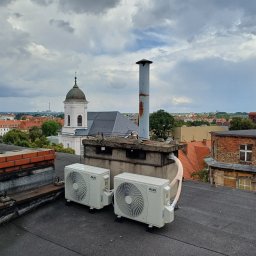OPTYMAL SYSTEM RAFAŁ KONDEJ - Dwa białe klimatyzatory AUX na dachu budynku z widokiem na miasto, kościół i kominy. Pochmurne niebo w tle.