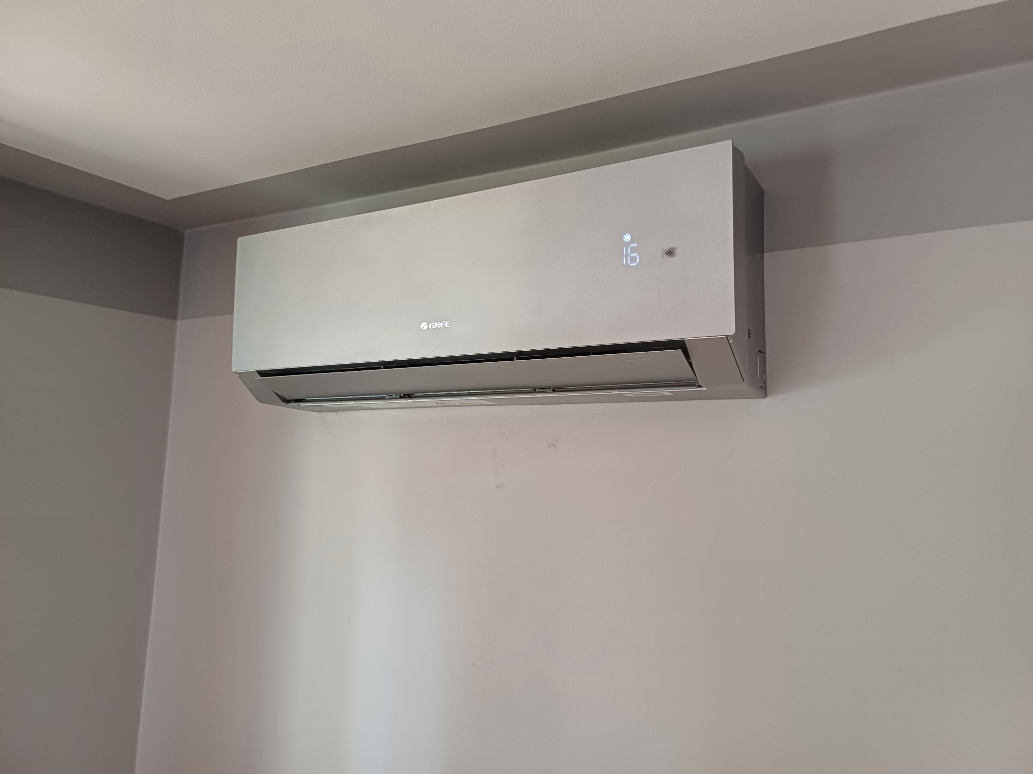 Szary klimatyzator Gree zamontowany na ścianie, wyświetlający temperaturę 16 stopni. Widoczne subtelne oświetlenie sufitowe.