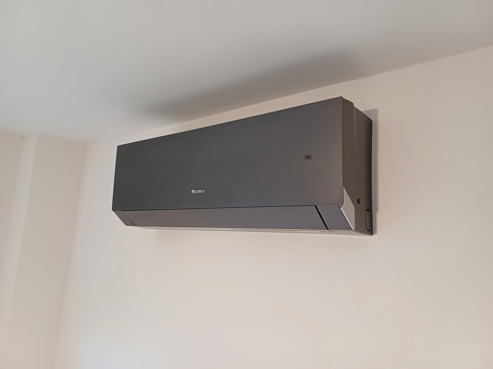 Czarny klimatyzator Gree zamontowany na jasnej ścianie. Minimalistyczny design, widoczne logo producenta, subtelne oświetlenie pomieszczenia.