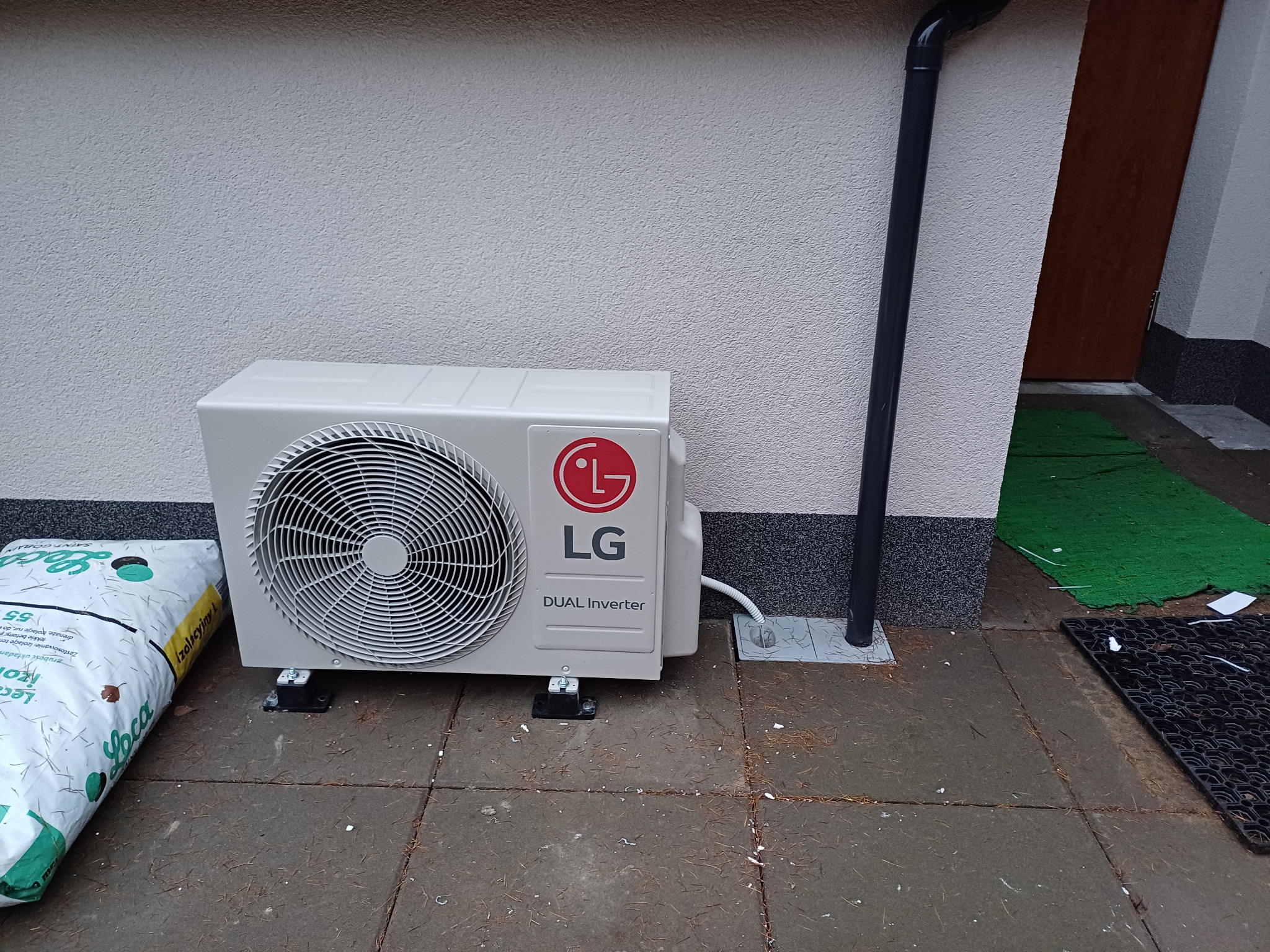 Biała zewnętrzna jednostka klimatyzacji LG Dual Inverter zamontowana na szarym bruku, obok rynny. W tle jasna ściana budynku i wejście.