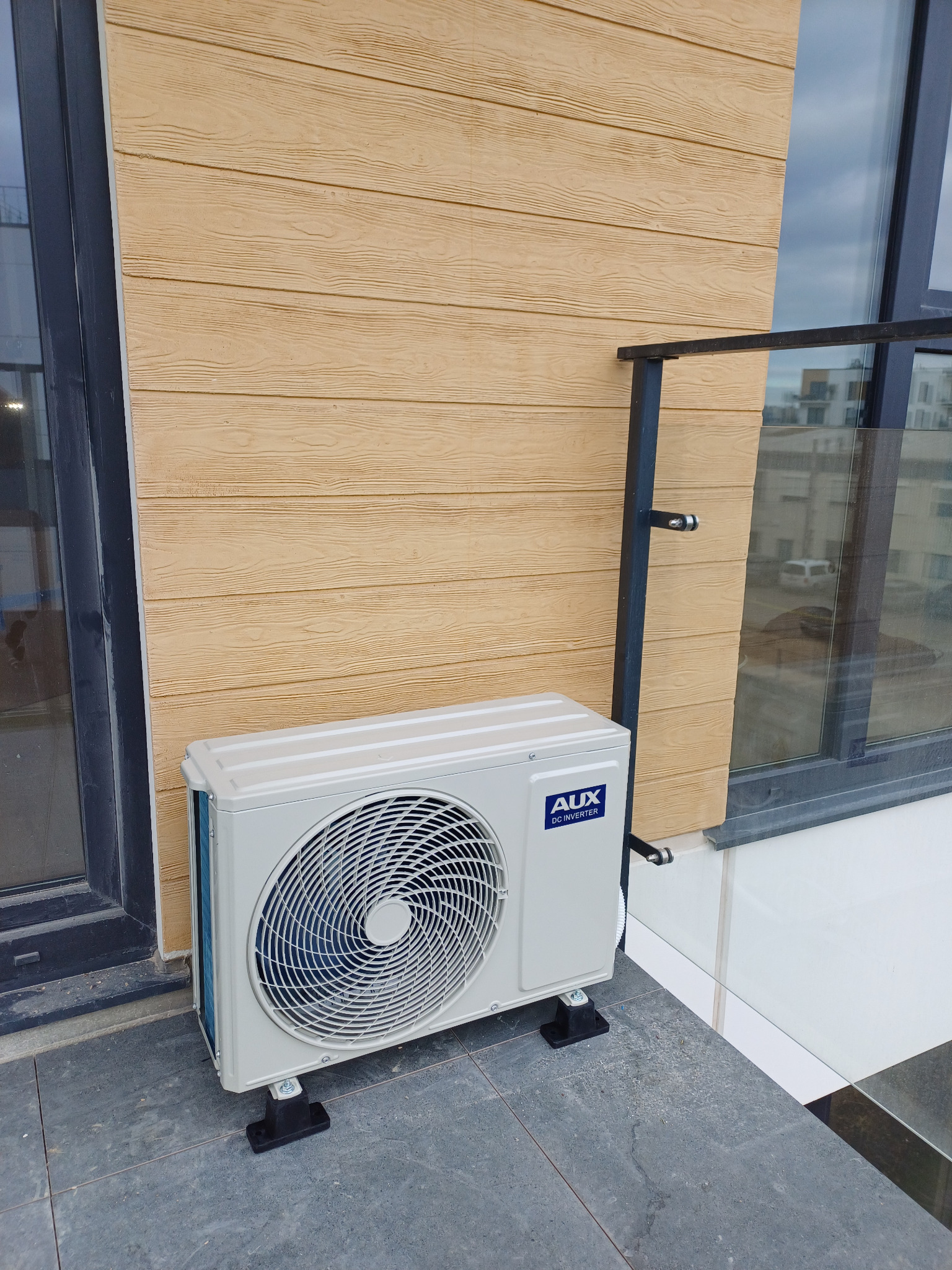 Zamontowana jednostka zewnętrzna klimatyzacji AUX na balkonie, widoczna na tle elewacji imitującej drewno i szklanej balustrady. Detal montażu na czarnych podkładkach.