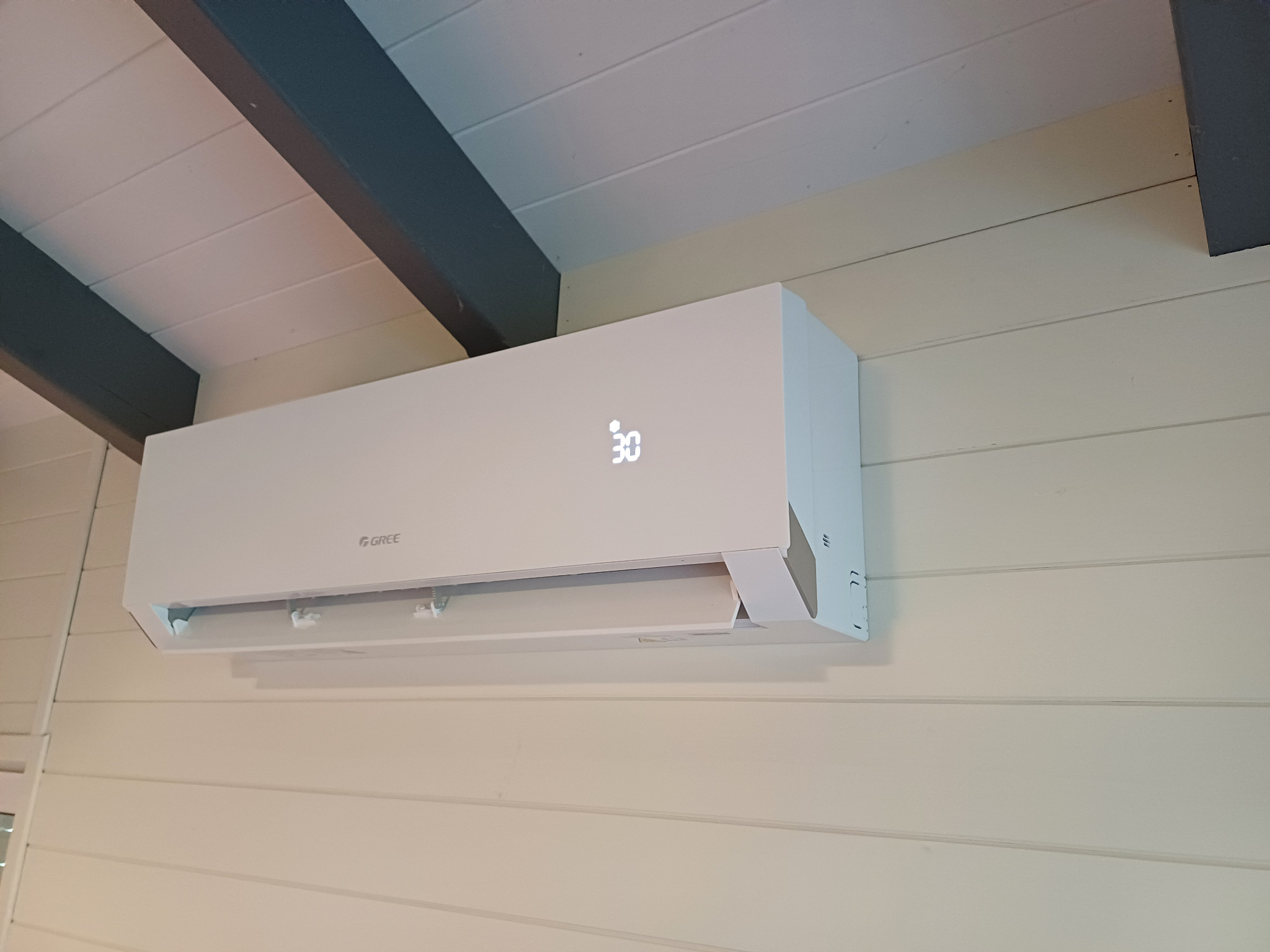 Biała klimatyzacja Gree na kremowej ścianie z drewnianą boazerią i ciemnymi belkami sufitowymi. Na wyświetlaczu widoczna temperatura 30 stopni.