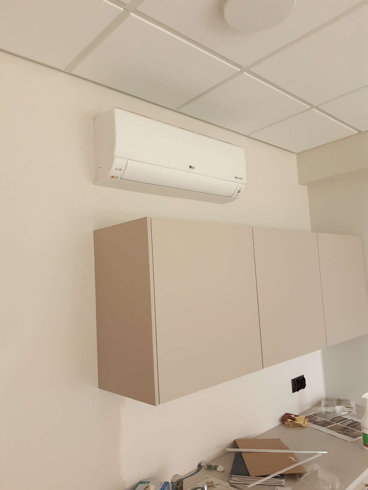 Biały klimatyzator LG zamontowany nad beżowymi szafkami w pomieszczeniu z podwieszanym sufitem. Widoczne gniazdko elektryczne i elementy wyposażenia wnętrza.