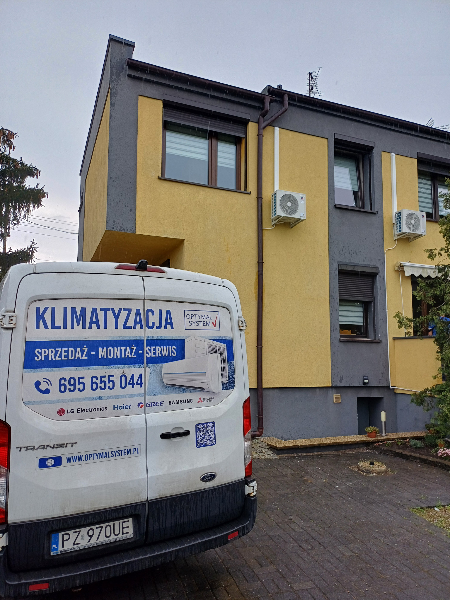 Żółto-szary budynek z zamontowanymi klimatyzatorami, widziany zza firmowego busa z reklamą sprzedaży, montażu i serwisu klimatyzacji. Widoczny numer telefonu.