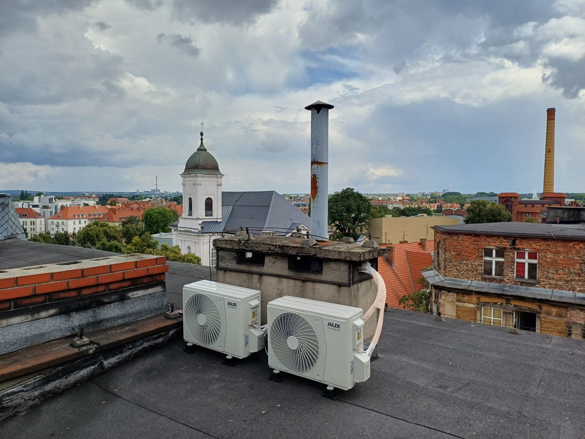 Dwa białe klimatyzatory AUX na dachu budynku z widokiem na miasto, kościół i kominy. Pochmurne niebo w tle.