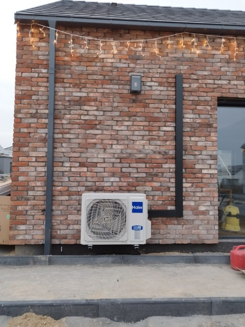 Biały klimatyzator Haier zamontowany na ceglanej ścianie domu z czarnymi rurami i świątecznymi lampkami, widok z zewnątrz.