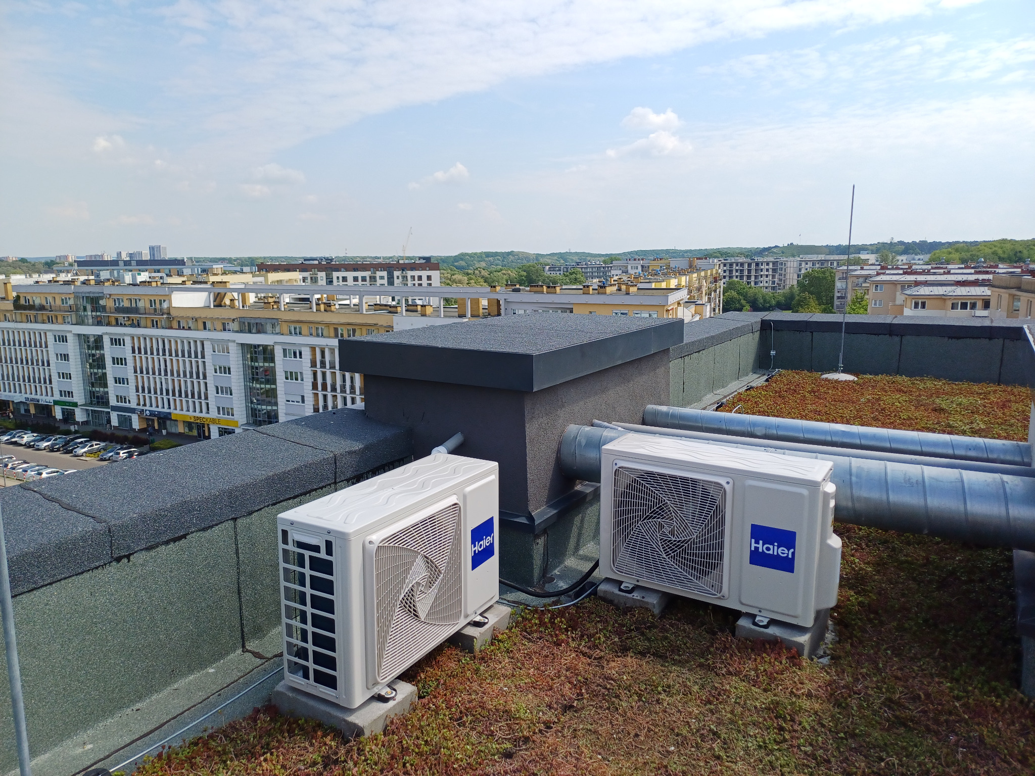 Dwa białe klimatyzatory Haier na dachu budynku z zieloną roślinnością, z widokiem na panoramę miasta i błękitne niebo w tle.