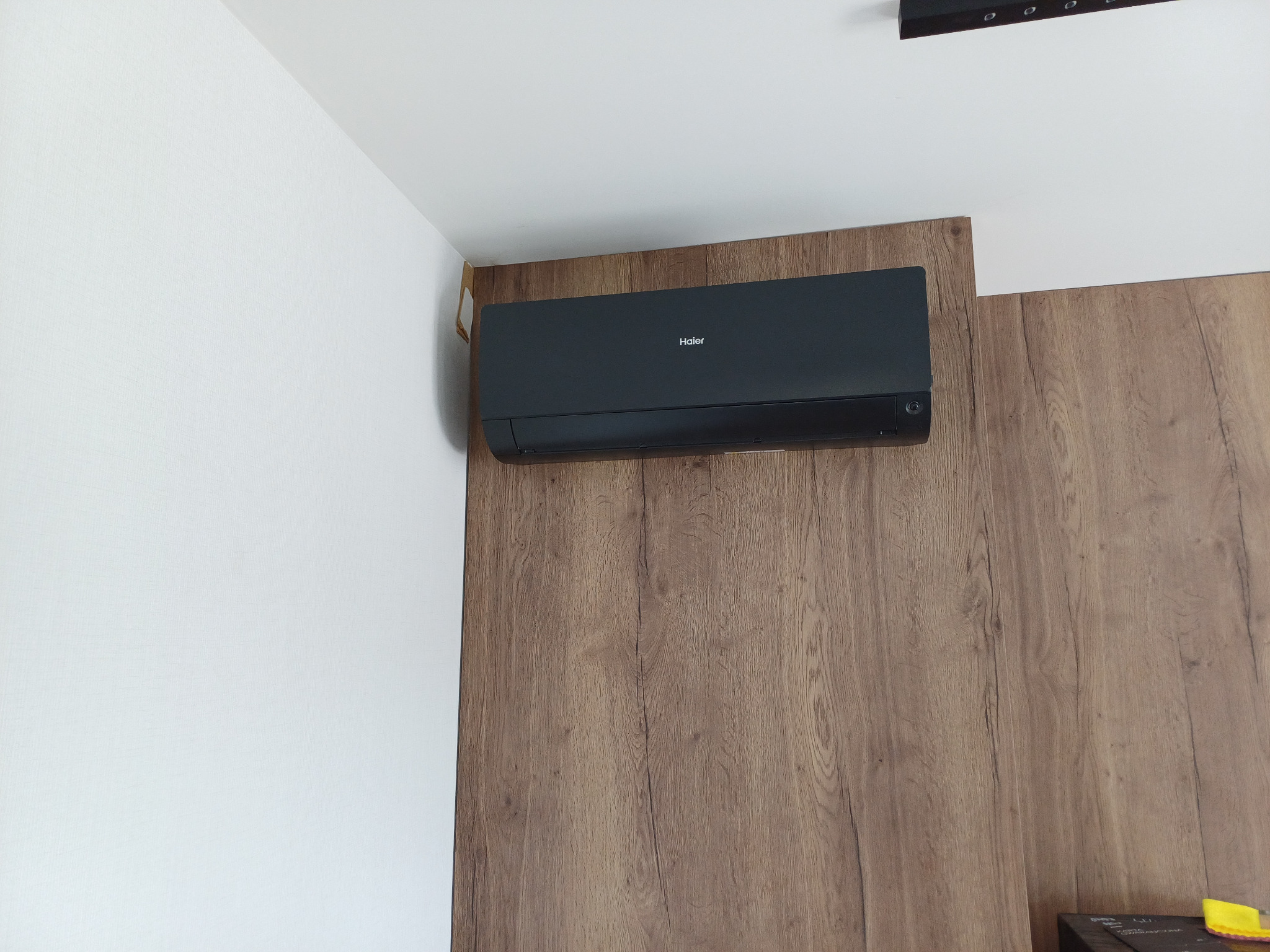 Czarny klimatyzator Haier na drewnianej ścianie, zamontowany w rogu pomieszczenia. Widoczny fragment sufitu z oświetleniem.