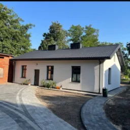 Biały dom po remoncie z szarym dachem i brukowanym podjazdem w słoneczny dzień. Widoczny budynek z czerwonej cegły obok.