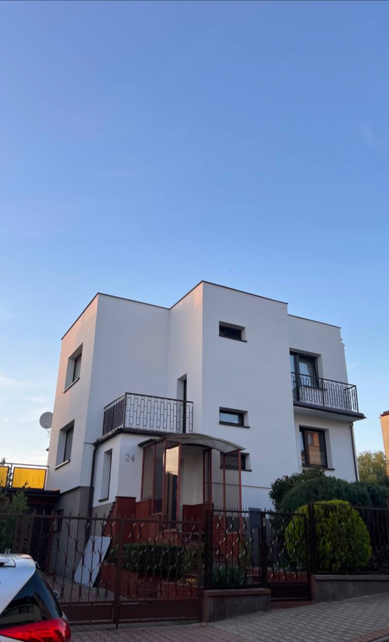 Biały, dwupiętrowy dom z balkonami i numerem 24, widziany z poziomu ulicy w pogodny dzień. Ogród i ogrodzenie przed domem.