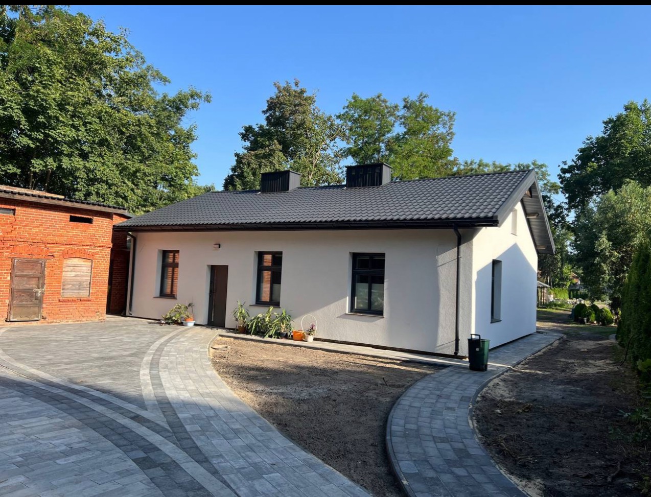 Biały dom po remoncie z szarym dachem i brukowanym podjazdem w słoneczny dzień. Widoczny budynek z czerwonej cegły obok.
