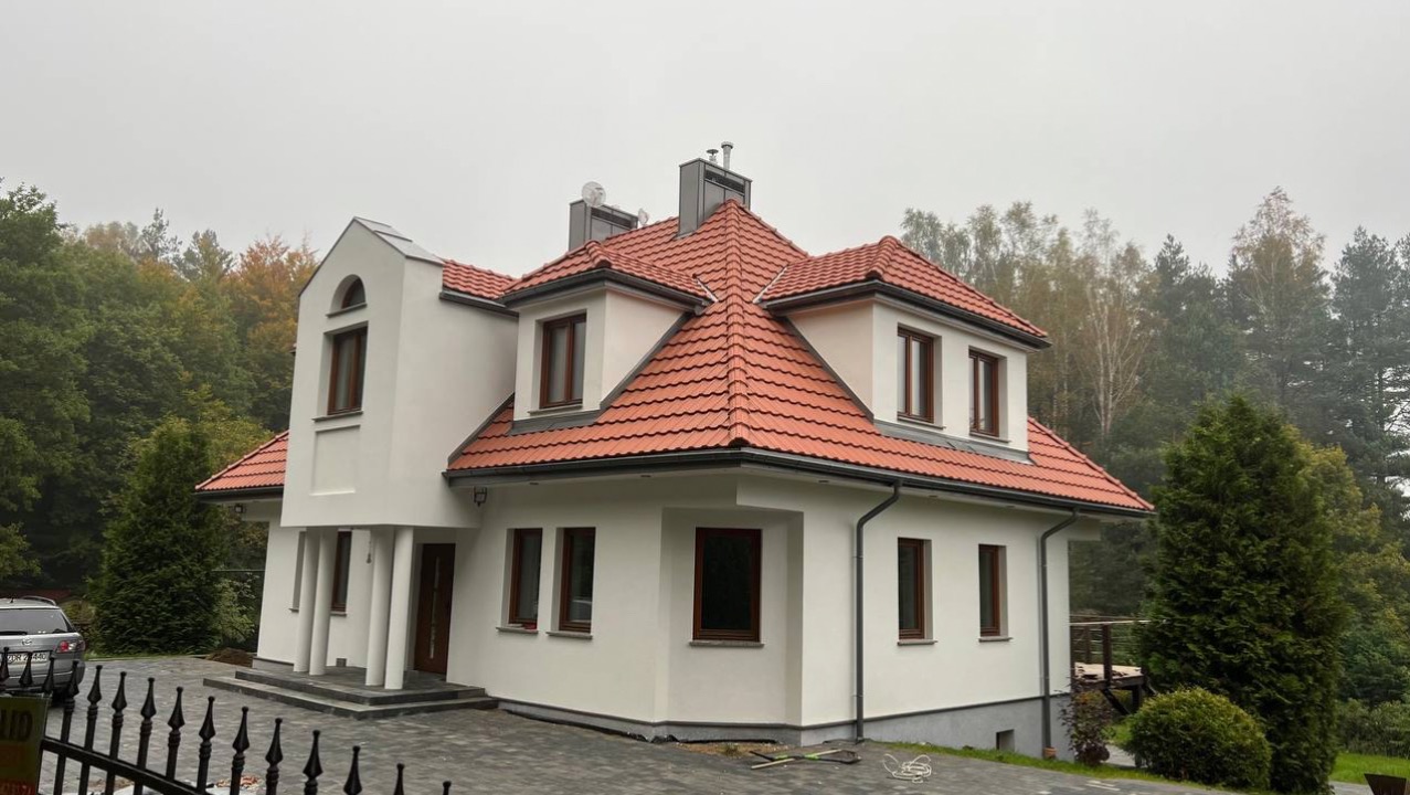 Elewacja domu jednorodzinnego po remoncie, z nową, czerwoną dachówką i świeżo pomalowaną, białą fasadą. Widok z poziomu chodnika w pochmurny dzień.