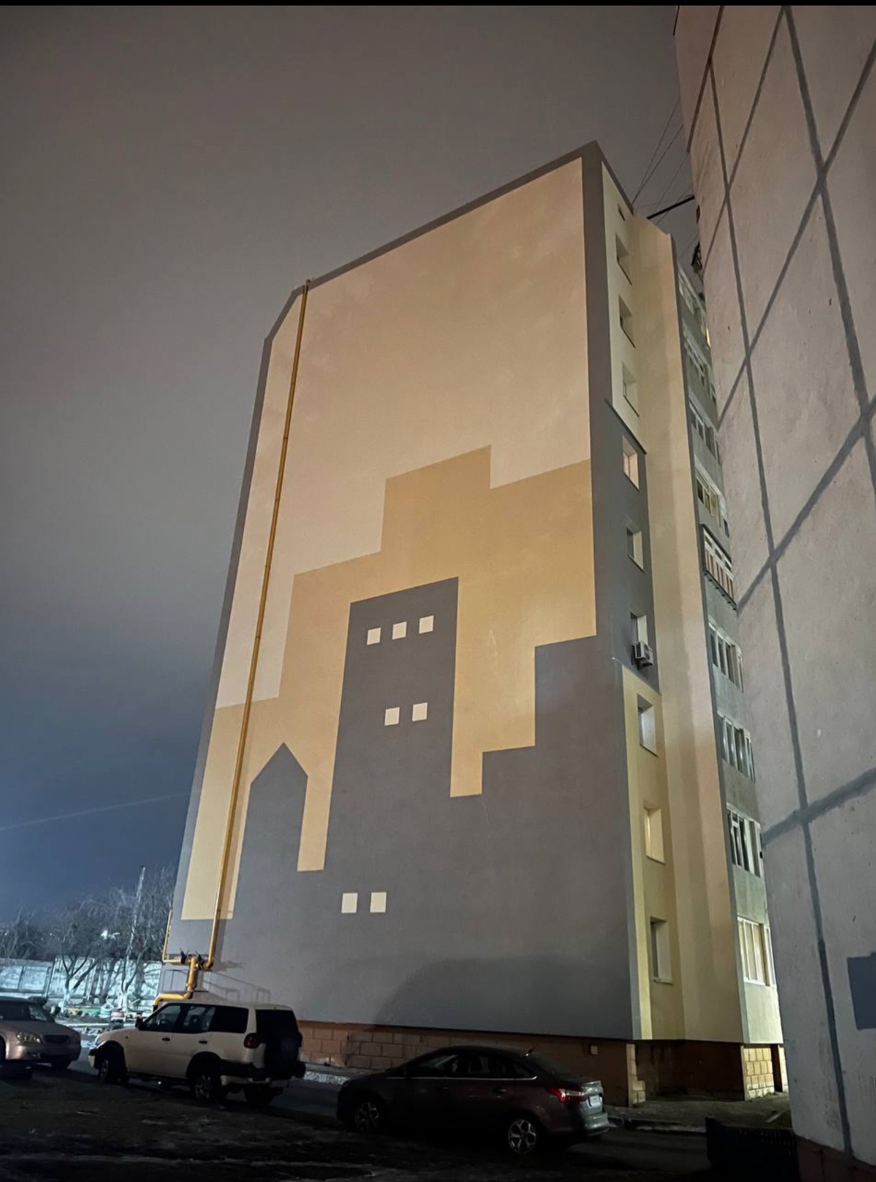 Wysoki budynek mieszkalny z abstrakcyjnym motywem miasta na ścianie, widoczny z dołu, nocne oświetlenie i zaparkowane samochody na pierwszym planie.