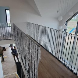 Szymon Klawikowski - Nowoczesne, czarne balustrady na schodach wewnątrz jasnego domu. Balustrady częściowo zabezpieczone folią ochronną, widoczni pracownicy.