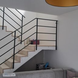 Szymon Klawikowski - Schody wewnętrzne z czarną, prostą balustradą. Widoczny fragment salonu z szarą kanapą. Proste, nowoczesne wykończenie.