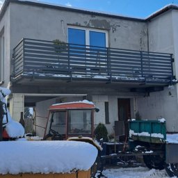 Szymon Klawikowski - Szara, pozioma, metalowa konstrukcja na balkonie dwukondygnacyjnego domu. Na pierwszym planie ciągniki rolnicze pokryte śniegiem.