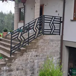 Nowoczesna balustrada schodowa z geometrycznym wzorem w Kartuzach. Czarna, metalowa konstrukcja na zewnątrz budynku, widok na zielony ogród i las.