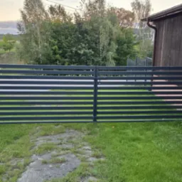 Nowoczesna, pozioma brama z metalowymi szczebelkami, prowadząca na posesję. Widok na zielony trawnik i budynek w tle, uchwycony w naturalnym oświetleniu.