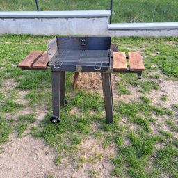 Szymon Klawikowski - Metalowy grill ogrodowy na kółkach z drewnianymi półkami, ustawiony na trawie przed betonowym ogrodzeniem. Widoczne spawane elementy konstrukcyjne.