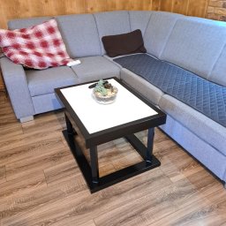 Szymon Klawikowski - Nowoczesny stolik kawowy z białym blatem i czarną ramą, na drewnianej podłodze, obok szarego narożnika z poduszkami. Wazonik z sukulentami na blacie.