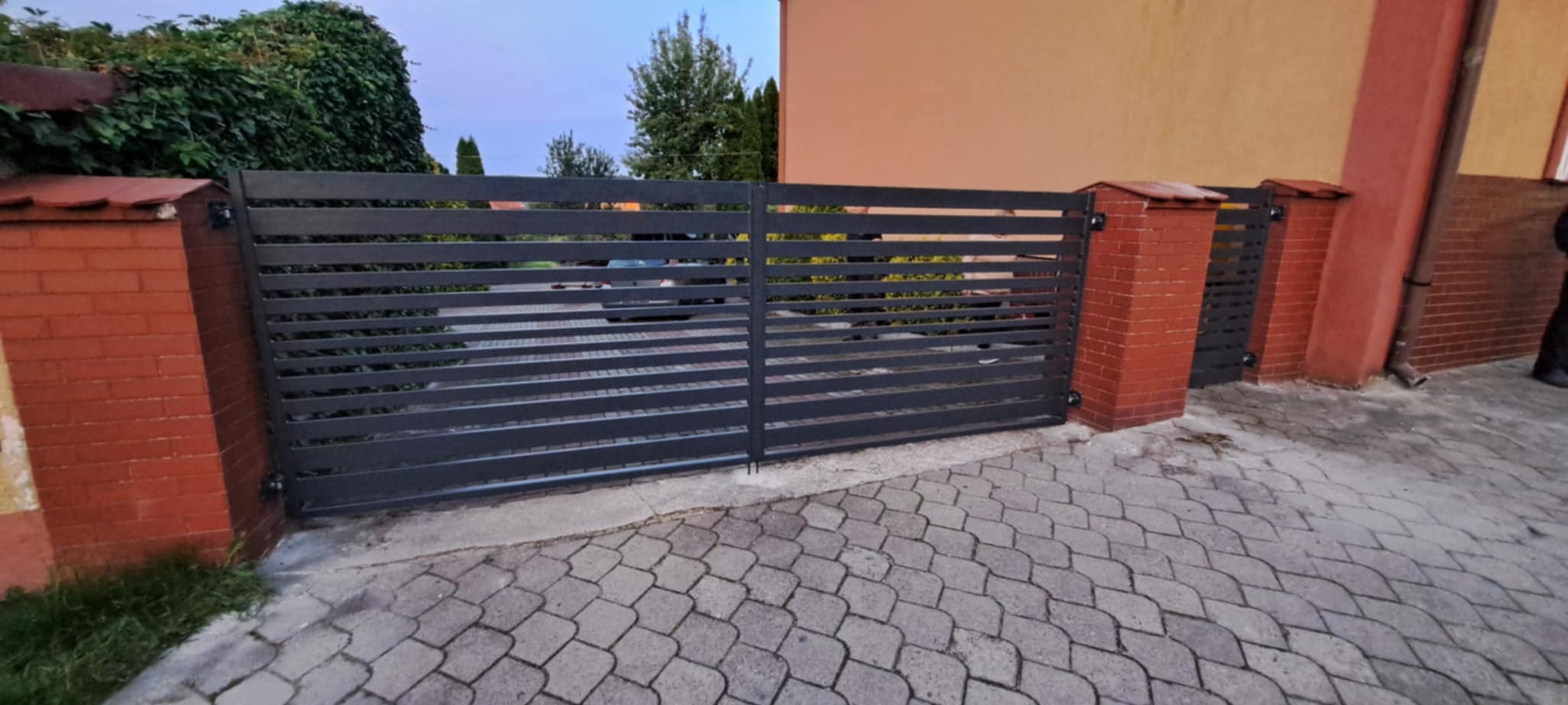 Nowoczesna, pozioma brama wjazdowa z ciemnego metalu, osadzona między ceglanymi słupkami. Widoczny fragment posesji i elewacji budynku w tle.