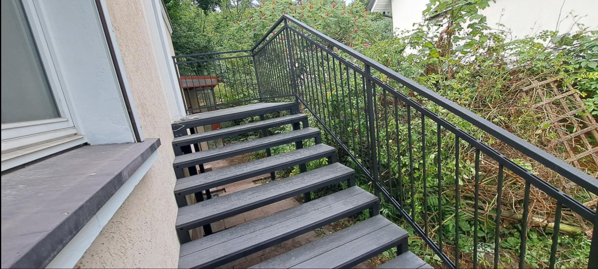 Zewnętrzne schody z metalową konstrukcją i poręczą, prowadzące do ogrodu. Widok z dołu, z boku budynku. Szare stopnie, czarna balustrada na tle zieleni.