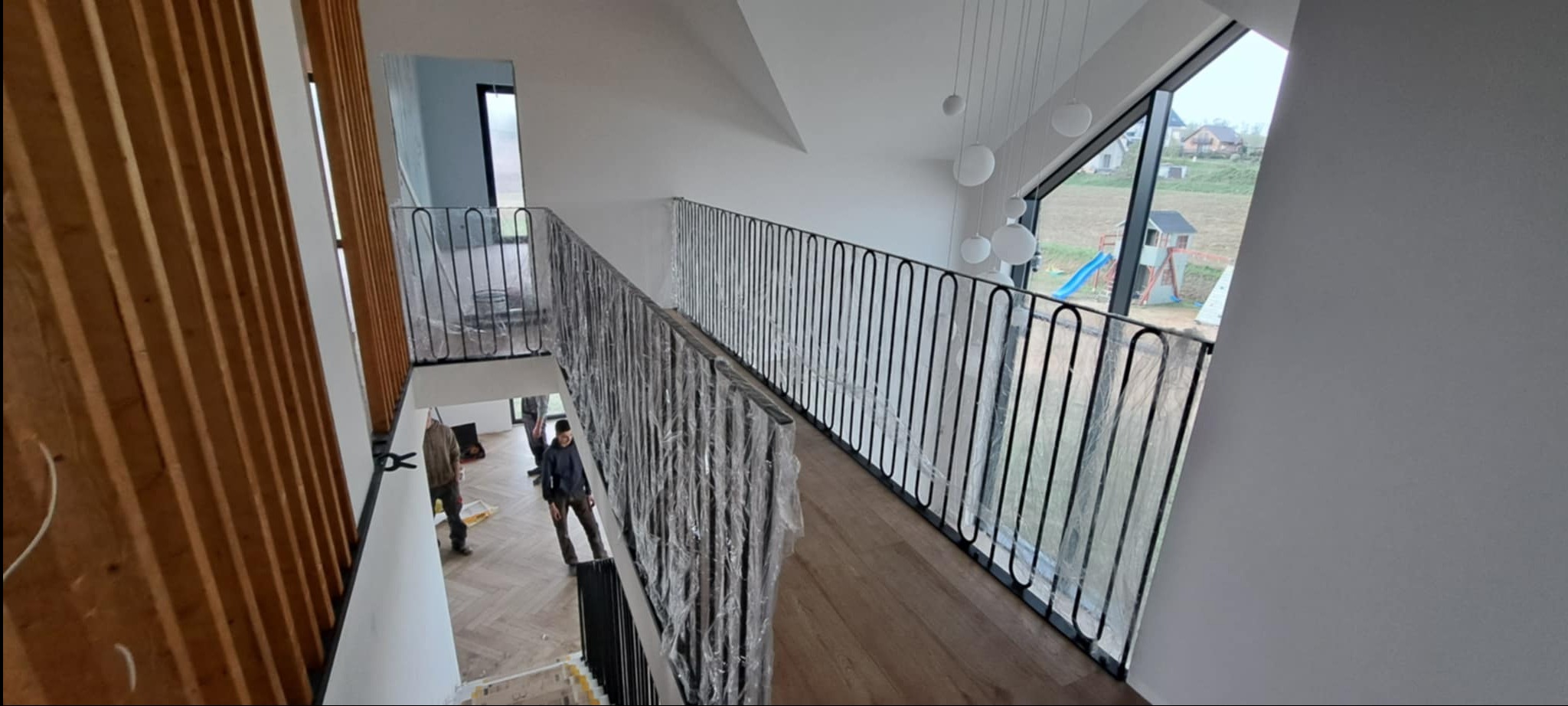 Nowoczesne, czarne balustrady na schodach wewnątrz jasnego domu. Balustrady częściowo zabezpieczone folią ochronną, widoczni pracownicy.