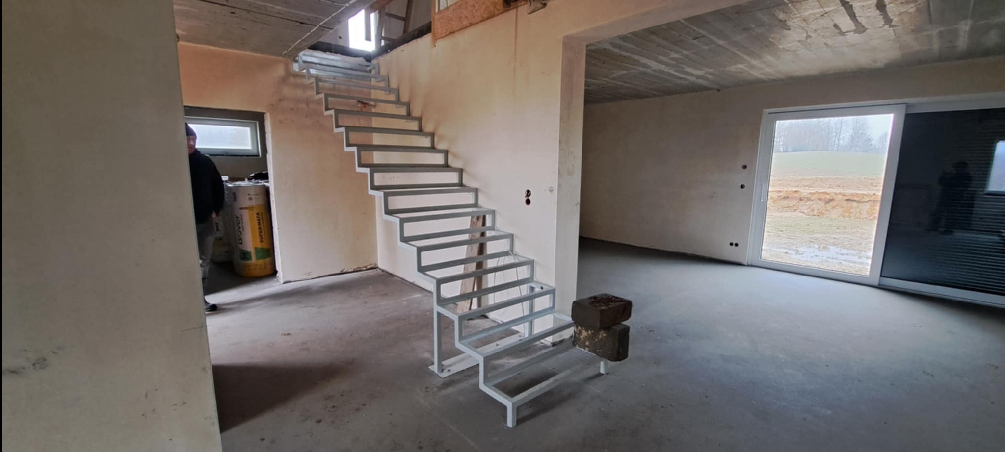 Surowe, metalowe schody modułowe w nowym domu, bez wykończenia. Widoczne cegły na podeście, surowy beton i duże okno przesuwne z widokiem na zewnątrz.