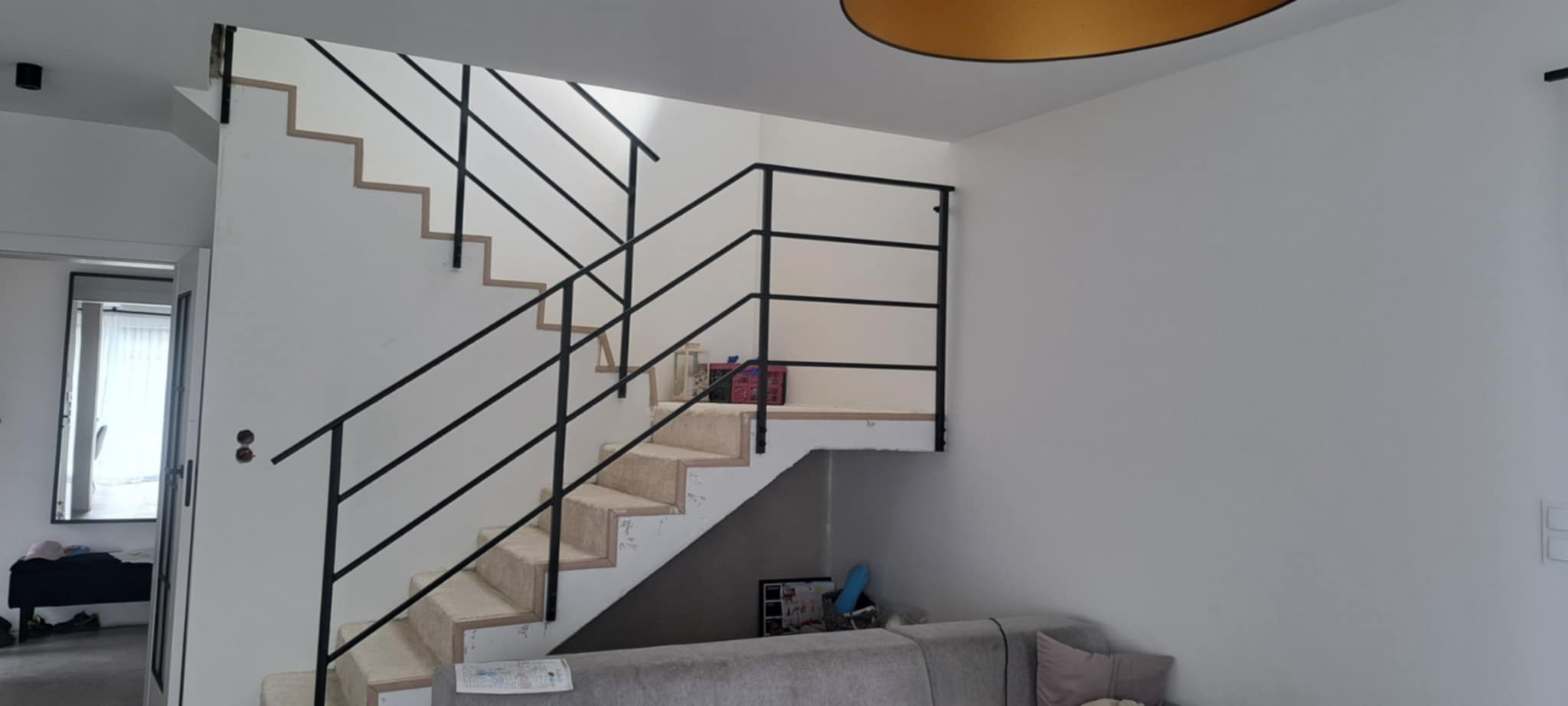 Schody wewnętrzne z czarną, prostą balustradą. Widoczny fragment salonu z szarą kanapą. Proste, nowoczesne wykończenie.