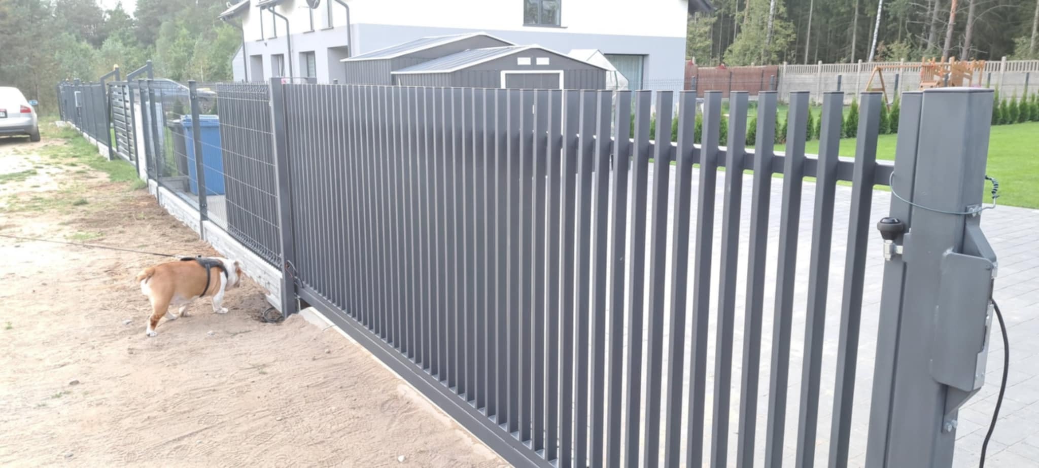 Nowoczesna, przesuwna brama wjazdowa z metalowymi sztachetami w kolorze szarym, z widocznym automatem. Pies rasy buldog francuski spaceruje wzdłuż ogrodzenia.