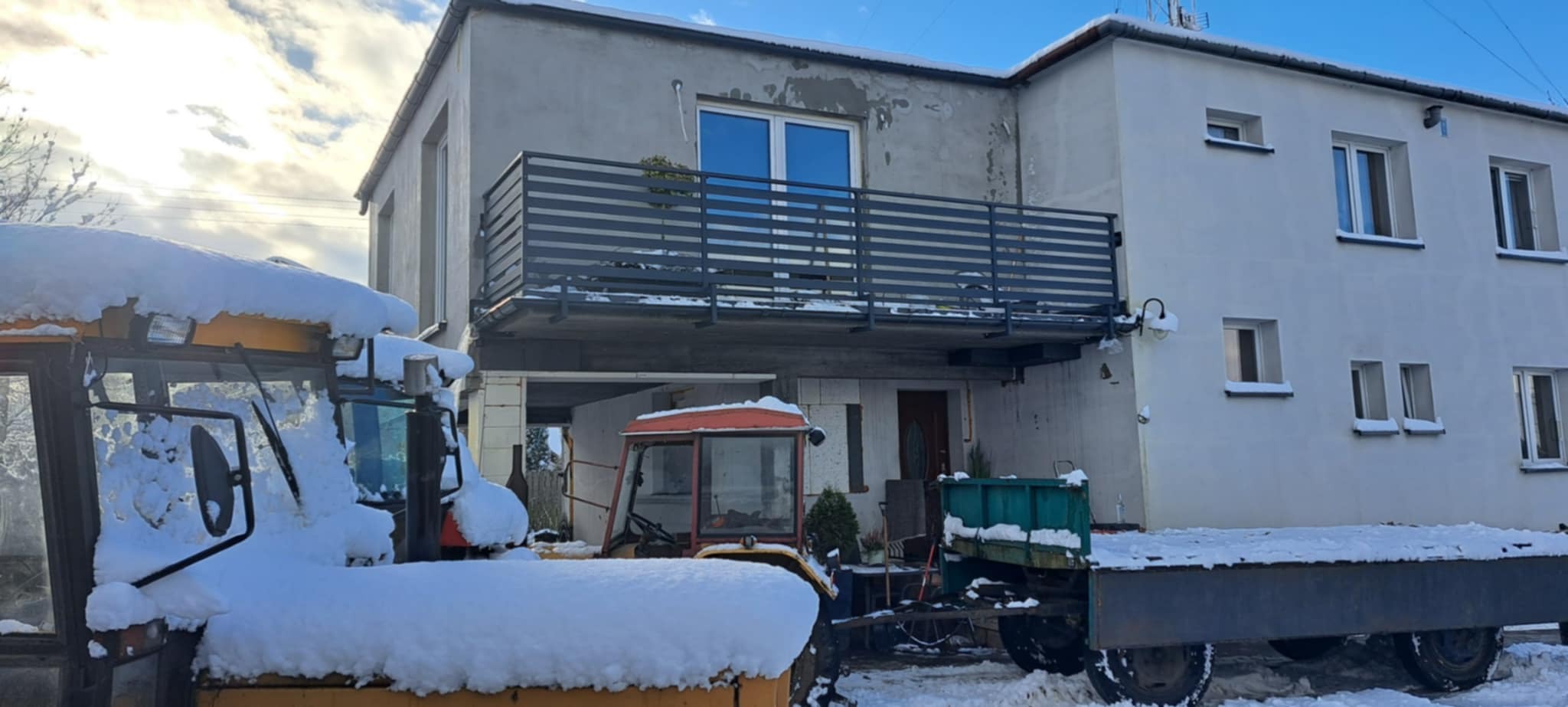 Szara, pozioma, metalowa konstrukcja na balkonie dwukondygnacyjnego domu. Na pierwszym planie ciągniki rolnicze pokryte śniegiem.