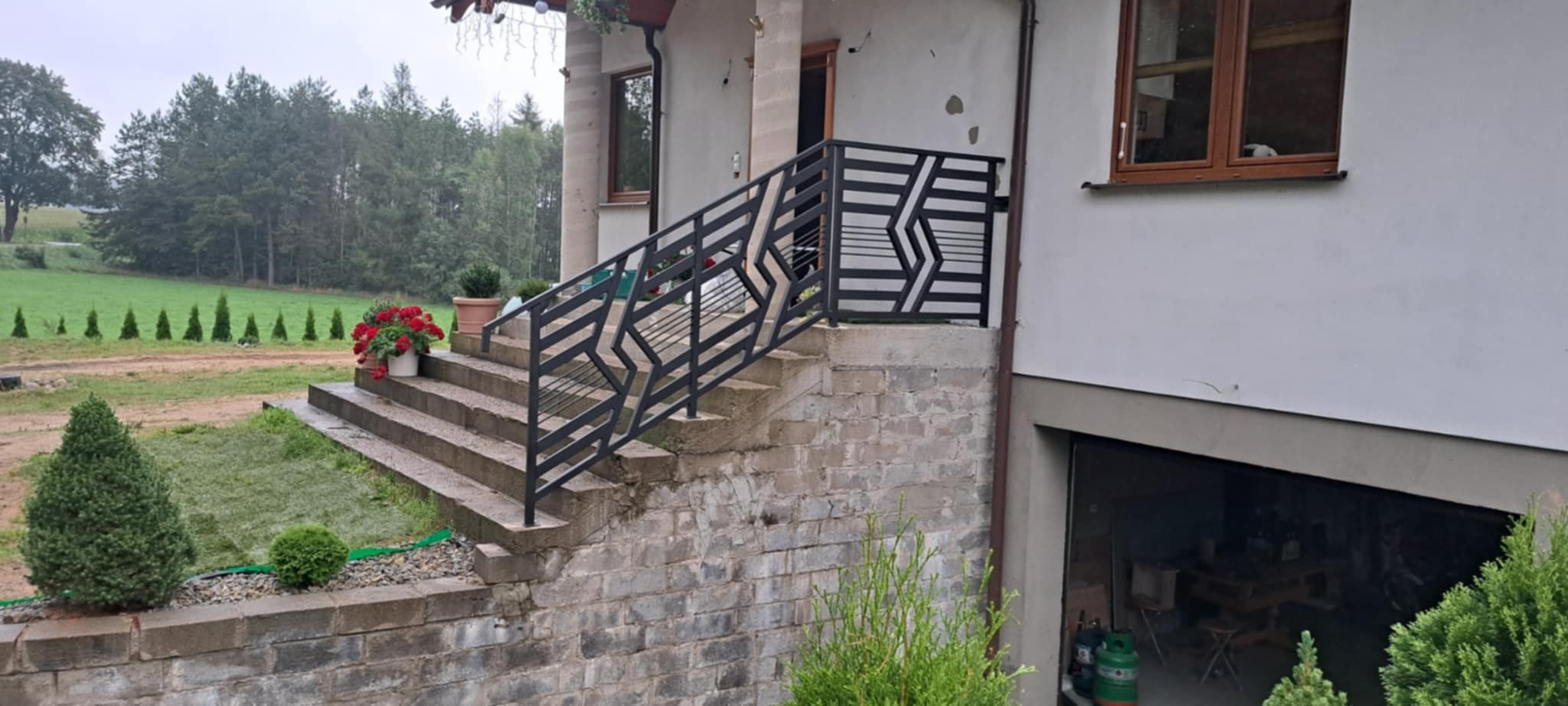 Nowoczesna balustrada schodowa z geometrycznym wzorem w Kartuzach. Czarna, metalowa konstrukcja na zewnątrz budynku, widok na zielony ogród i las.