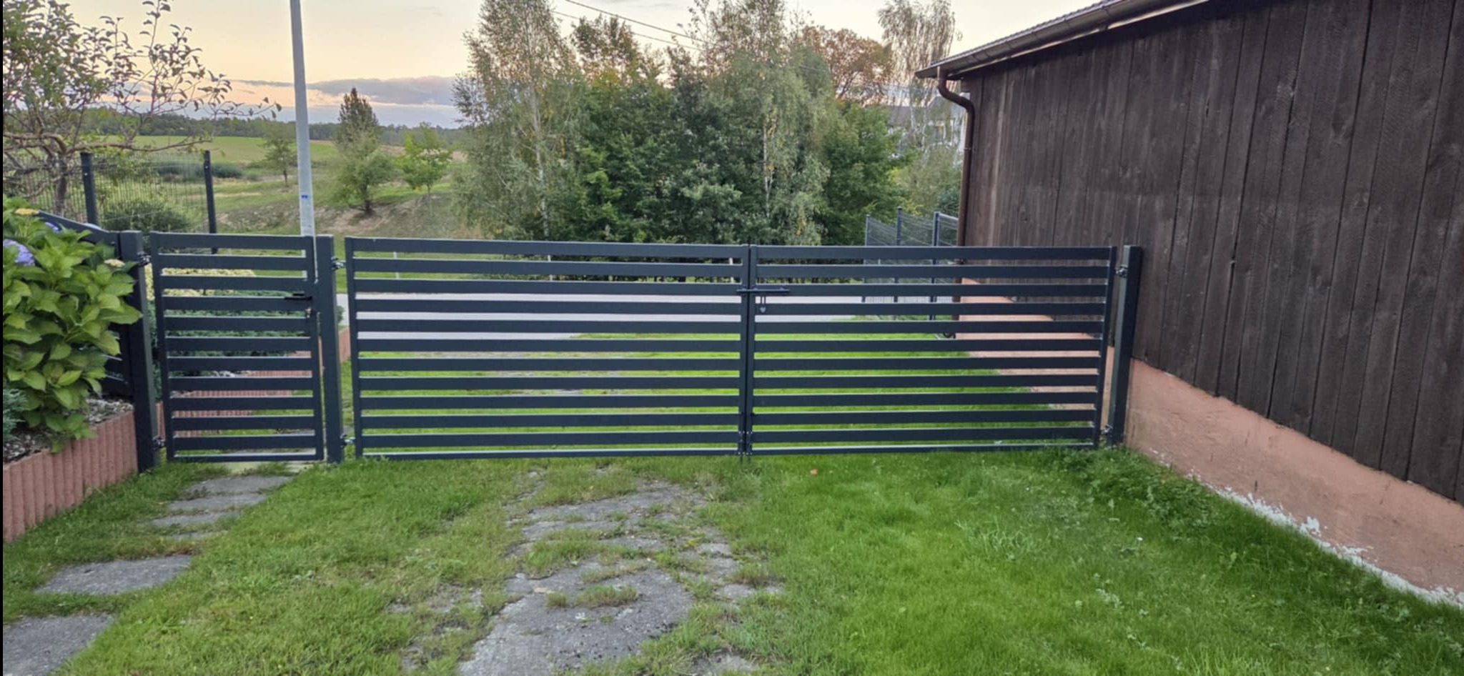 Nowoczesna, pozioma brama z metalowymi szczebelkami, prowadząca na posesję. Widok na zielony trawnik i budynek w tle, uchwycony w naturalnym oświetleniu.